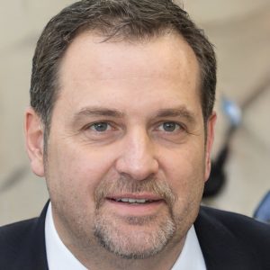 Portrait of a smiling man with short hair and a suit, looking at the camera with a neutral background.