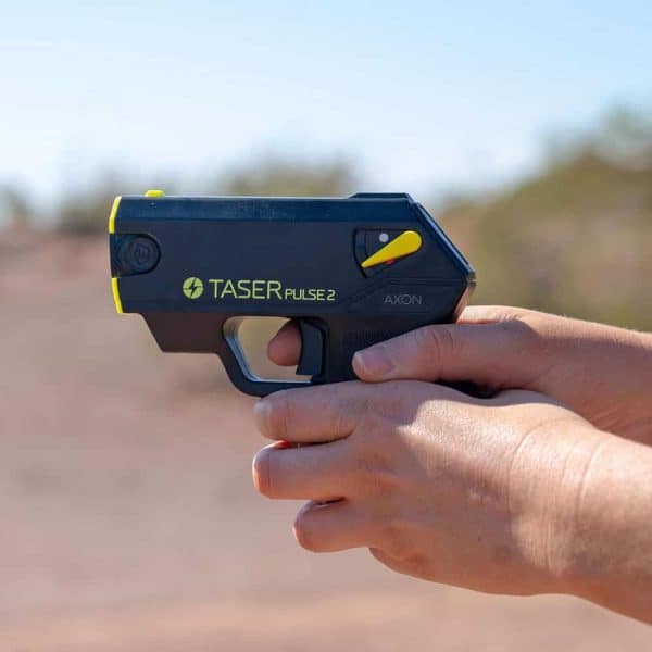 Hands holding Taser Pulse 2 against a blurred outdoor background, focusing on its design and detail.