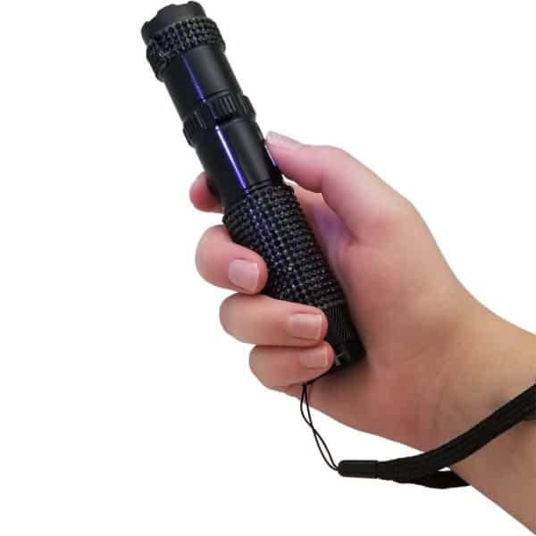 Hand holding a small black flashlight with textured grip and wrist strap, against a white background.