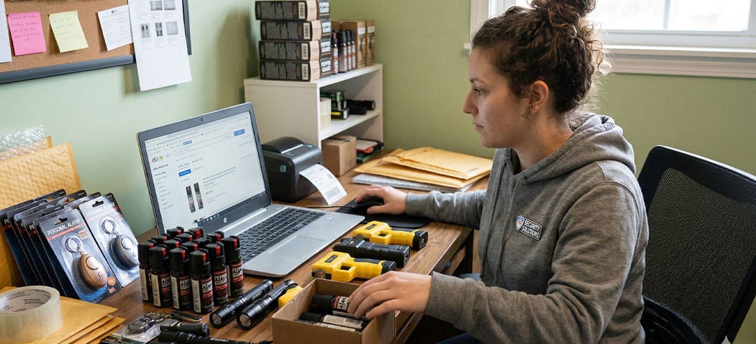 Person working at a desk with pepper spray products and a laptop, organizing items for online sales.
