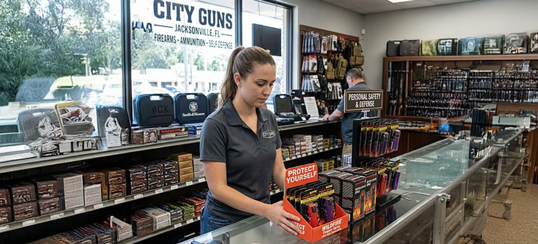 Employee organizing pepper spray display at City Guns in Jacksonville. Firearms and self-defense items visible in store.